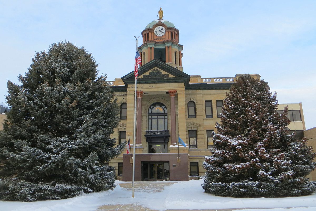 The Brown County Courthouse in Aberdeen, a tan stone building with a clock tower and green dome, between two snow-dusted spruce trees in winter