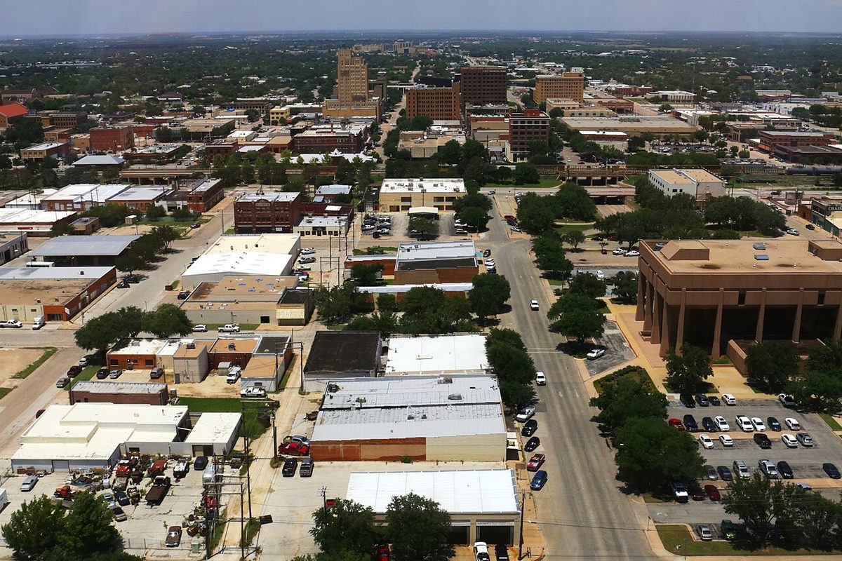 Aerial view over downtown Abilene on a hazy summer day, with flat-roofed buildings, a tree-lined street of parked cars and taller brick towers toward the horizon