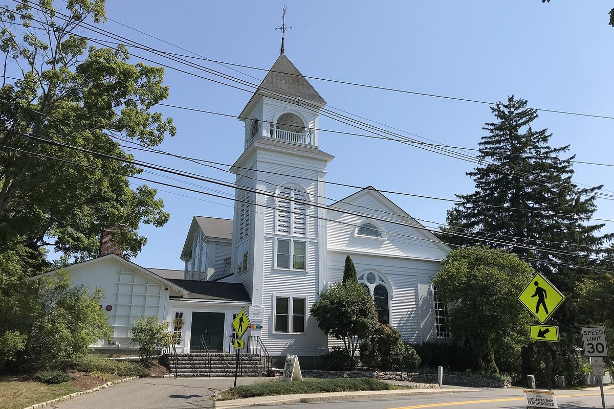 The white clapboard Acton Congregational Church with its tall steeple and weathervane, behind a pedestrian crossing sign and a 30 mph speed limit sign