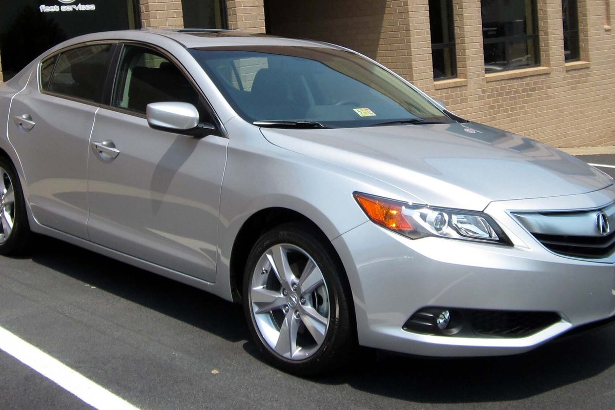 A silver 2013 Acura ILX sedan parked in front of a brick building, seen from the front passenger side