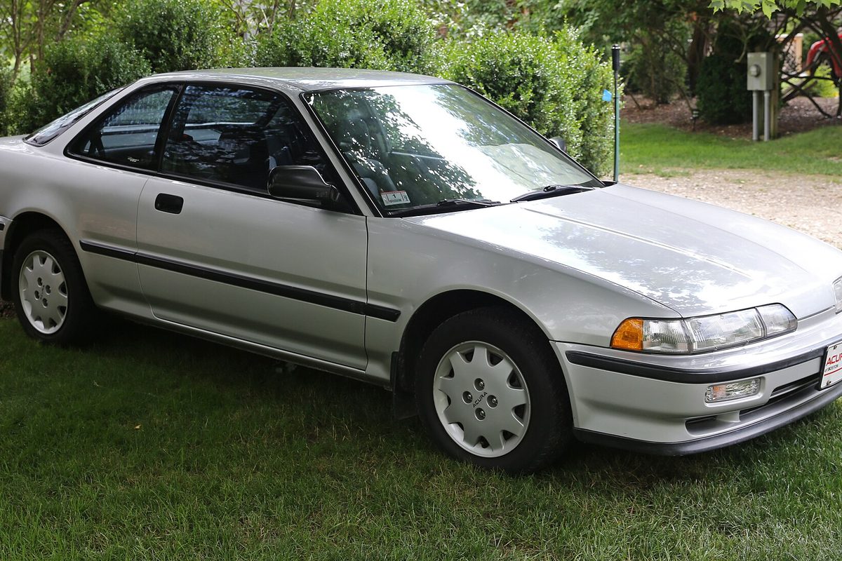 A silver second-generation Acura Integra three-door hatchback parked on a green lawn in front of hedges