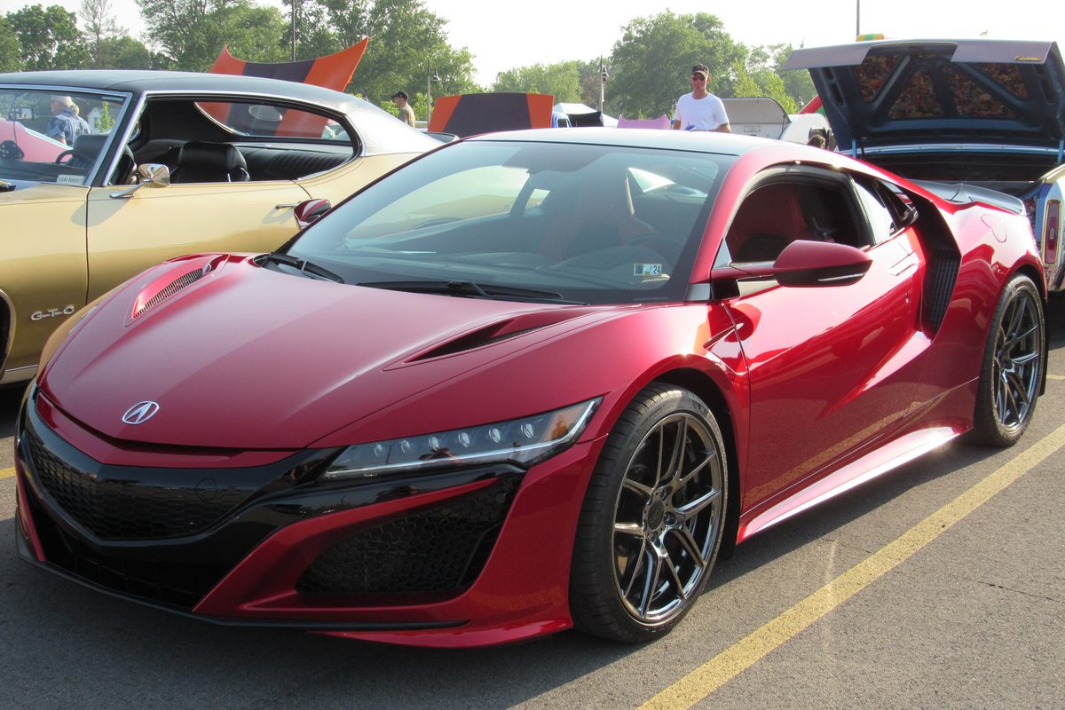 A red second-generation Acura NSX coupe with black wheels at an outdoor car show, an older gold car parked beside it