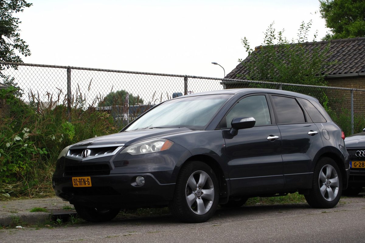 A dark gray first-generation Acura RDX parked on a street beside a chain-link fence and tall weeds