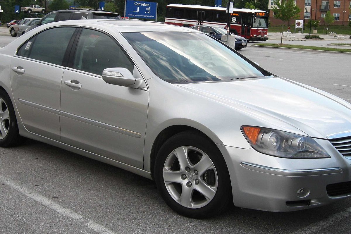 A silver 2005-2008 Acura RL sedan parked in a shopping lot, with a red bus passing behind it