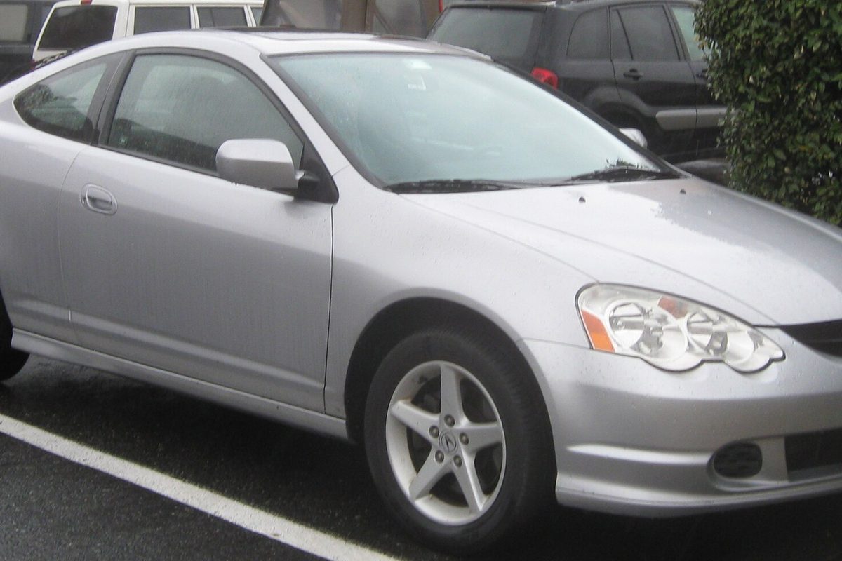 A silver 2002-2004 Acura RSX hatchback coupe parked on wet pavement beside a hedge