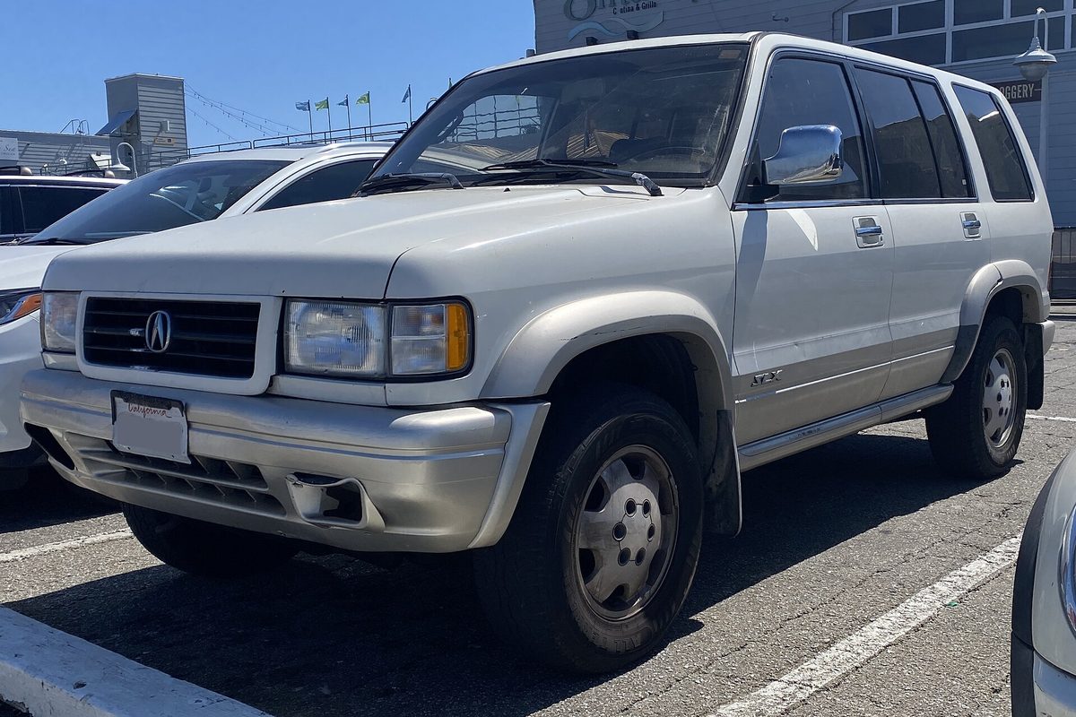 A cream-white and silver Acura SLX SUV with steel wheels, parked in a waterfront lot under a clear blue sky
