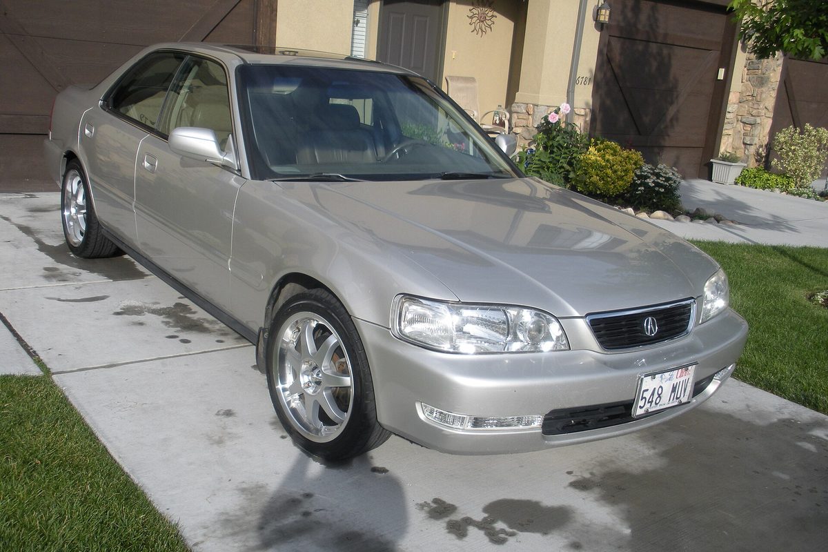 A champagne-silver first-generation Acura TL sedan with aftermarket alloy wheels, parked on a concrete driveway in front of a garage
