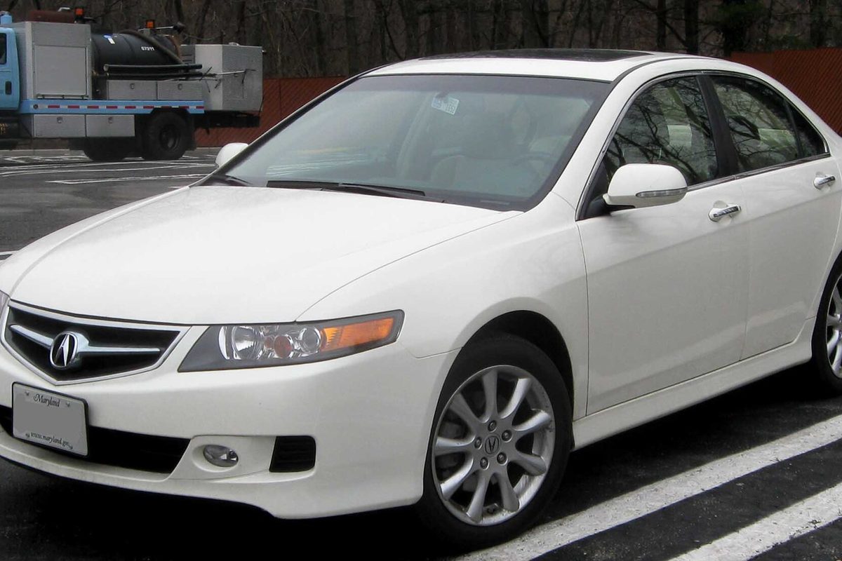 A white first-generation Acura TSX sedan with a sunroof, parked in a lot with a utility truck behind it