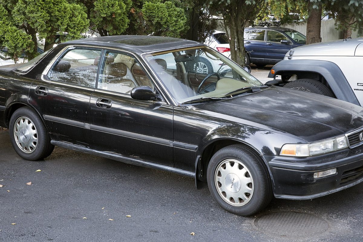 A black Acura Vigor sedan with faded paint and plain silver hubcaps, parked on a street beside green shrubs