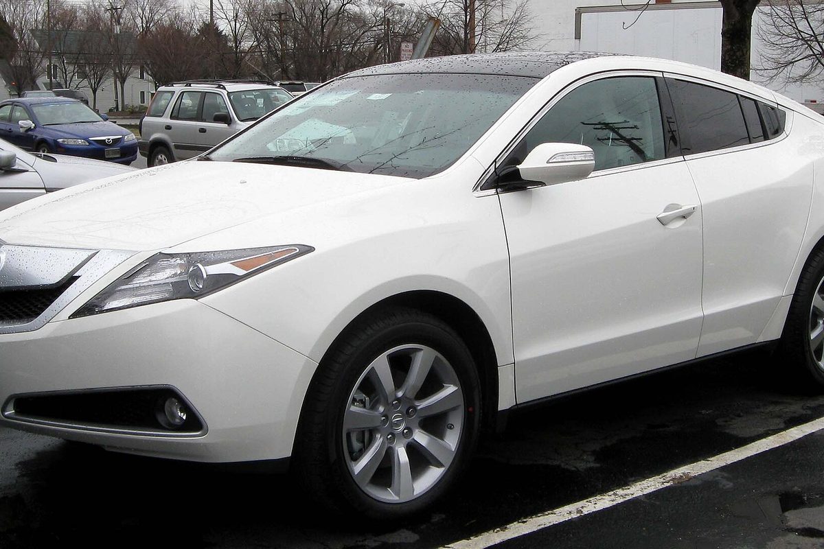 A white first-generation Acura ZDX crossover seen from the front left, parked on wet pavement with bare trees behind it