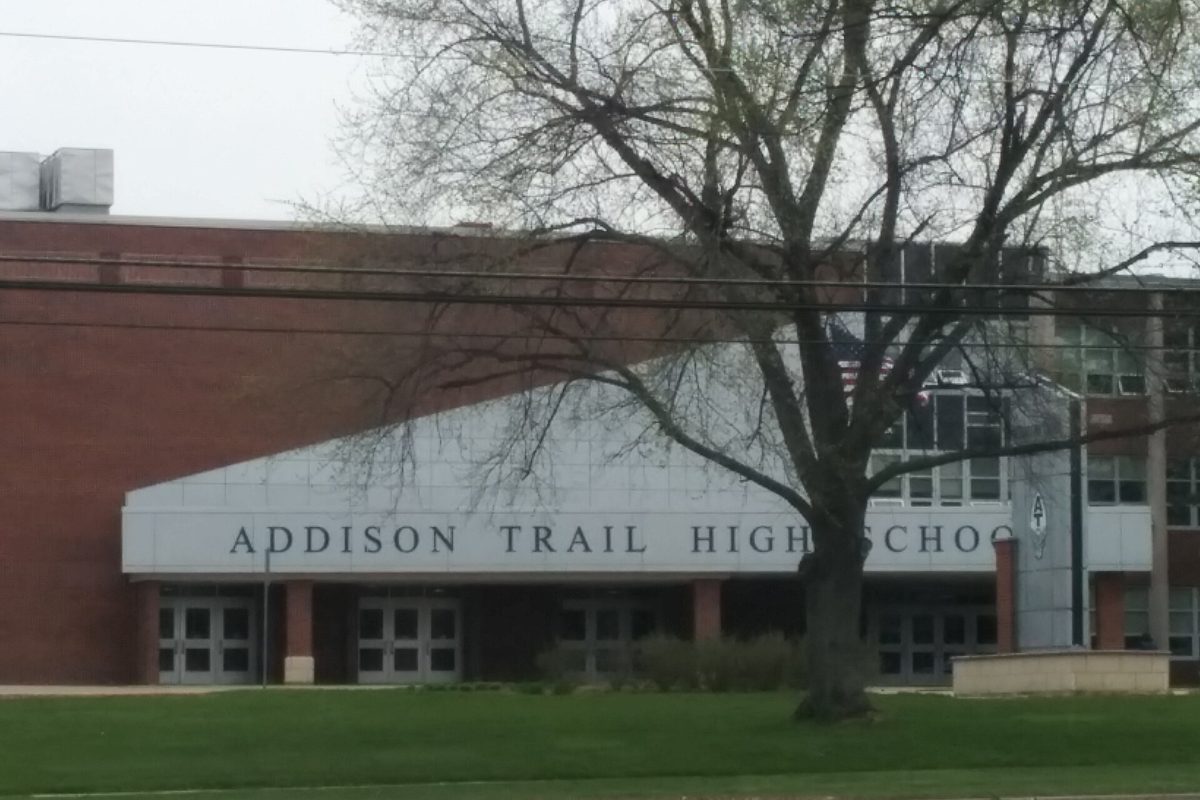 The front of Addison Trail High School, a brick and white panel building with its name in large letters, behind a bare tree and a lawn
