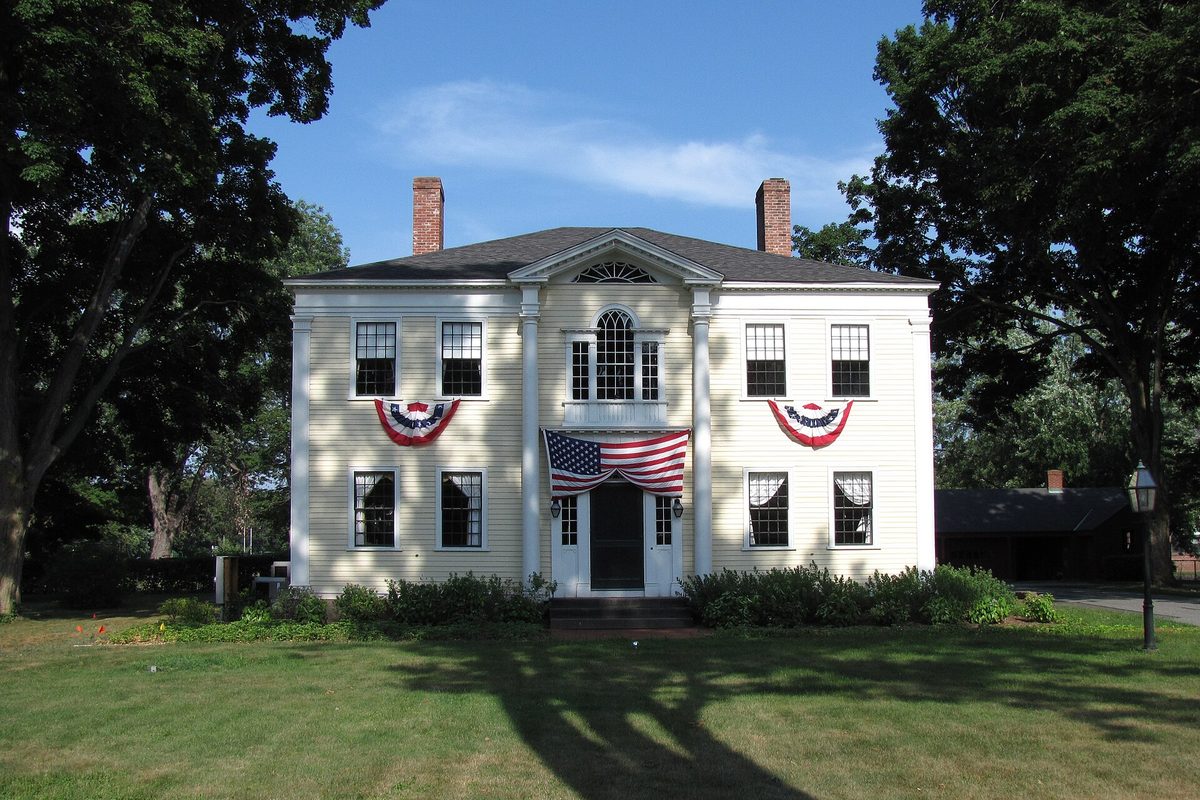 The Capt. Charles Leonard House in Agawam, a white two-story house with twin brick chimneys and red, white and blue bunting, under tall shade trees