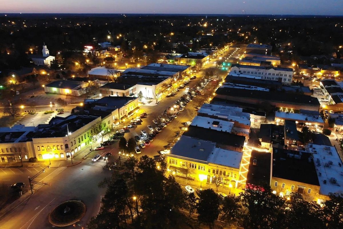Downtown Aiken from a drone at twilight, with lit storefronts and parked cars lining a wide main street