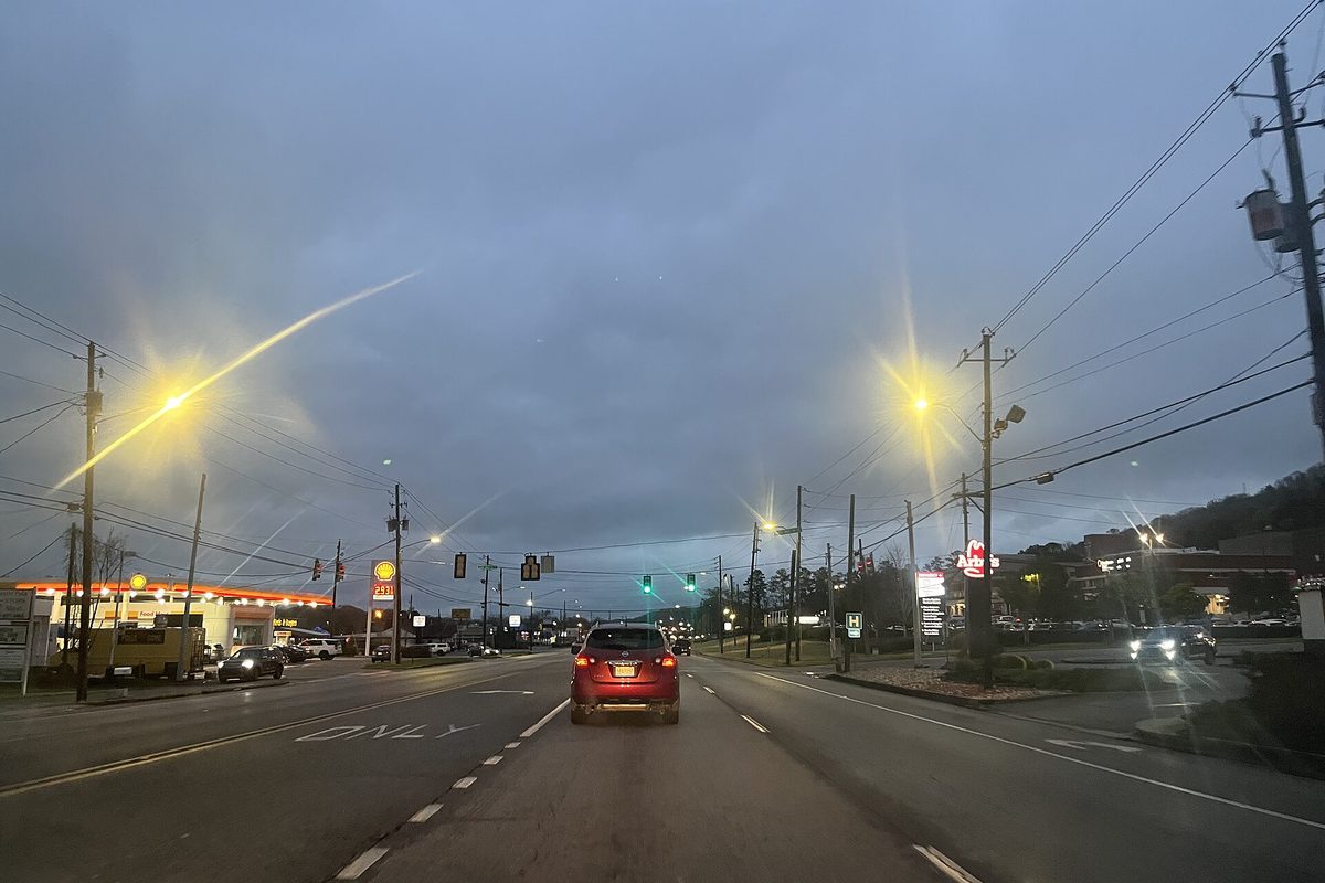 A four-lane road through downtown Alabaster at dusk, a red SUV in the near lane, streetlights glowing and a gas station canopy on the left