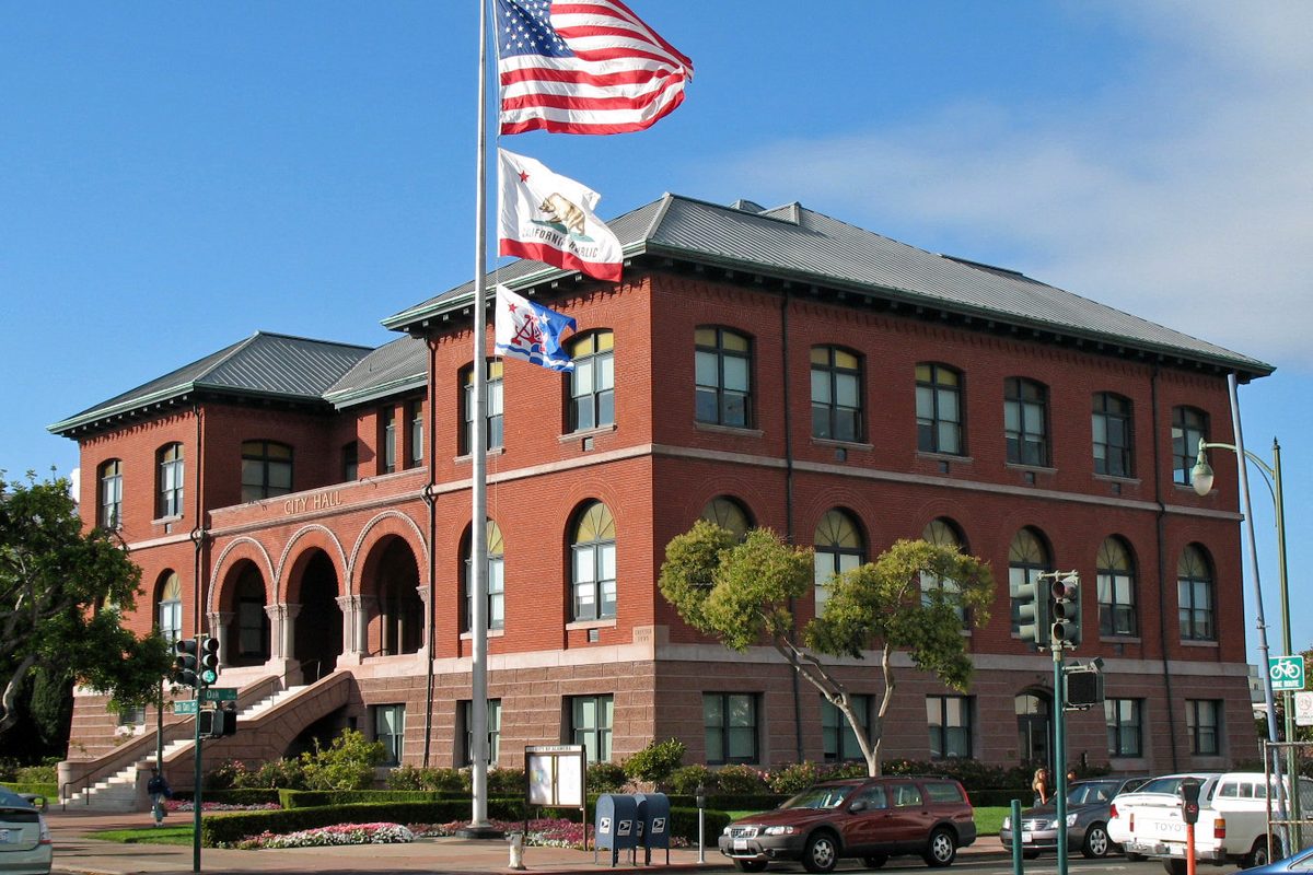 Alameda City Hall, a red brick building with arched windows and a slate roof, with U.S., California and city flags flying out front on a sunny day