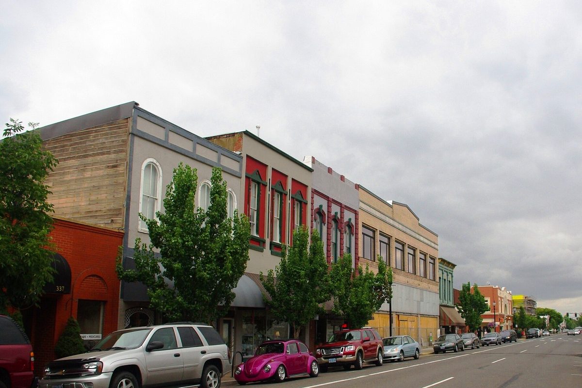 Two-story brick and painted storefronts on a downtown Albany street, with a pink Volkswagen Beetle and SUVs parked at the curb under a gray sky
