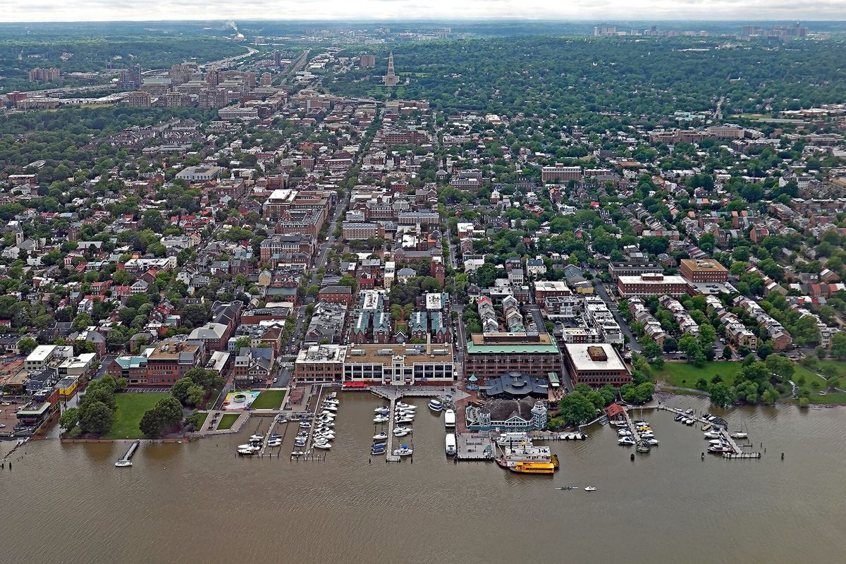 Aerial view of Old Town Alexandria, with dense blocks of brick rowhouses and leafy streets running up from a riverfront marina and a tall tower on the horizon