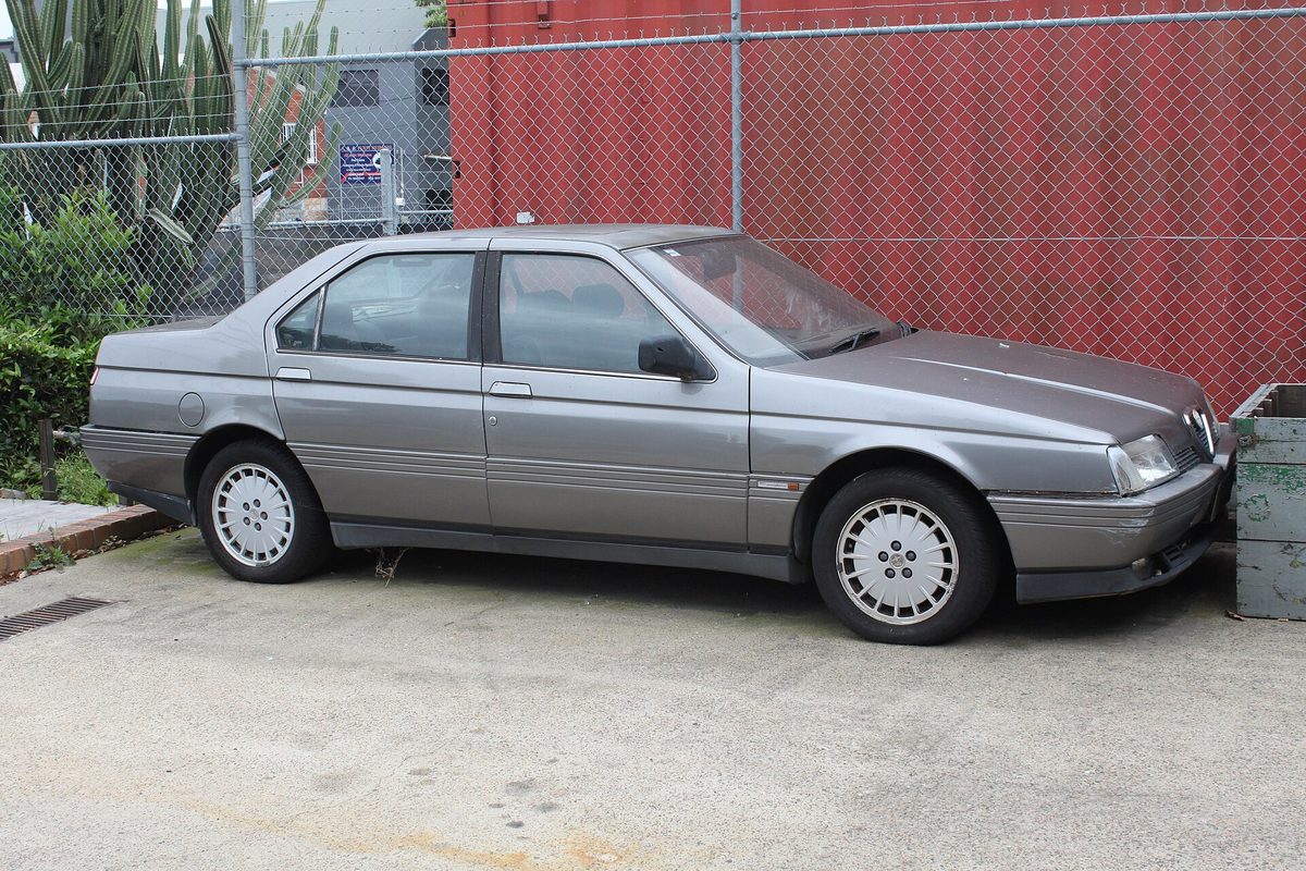 A grey Alfa Romeo 164 four-door sedan with dished alloy wheels, parked beside a chain-link fence and a red shipping container