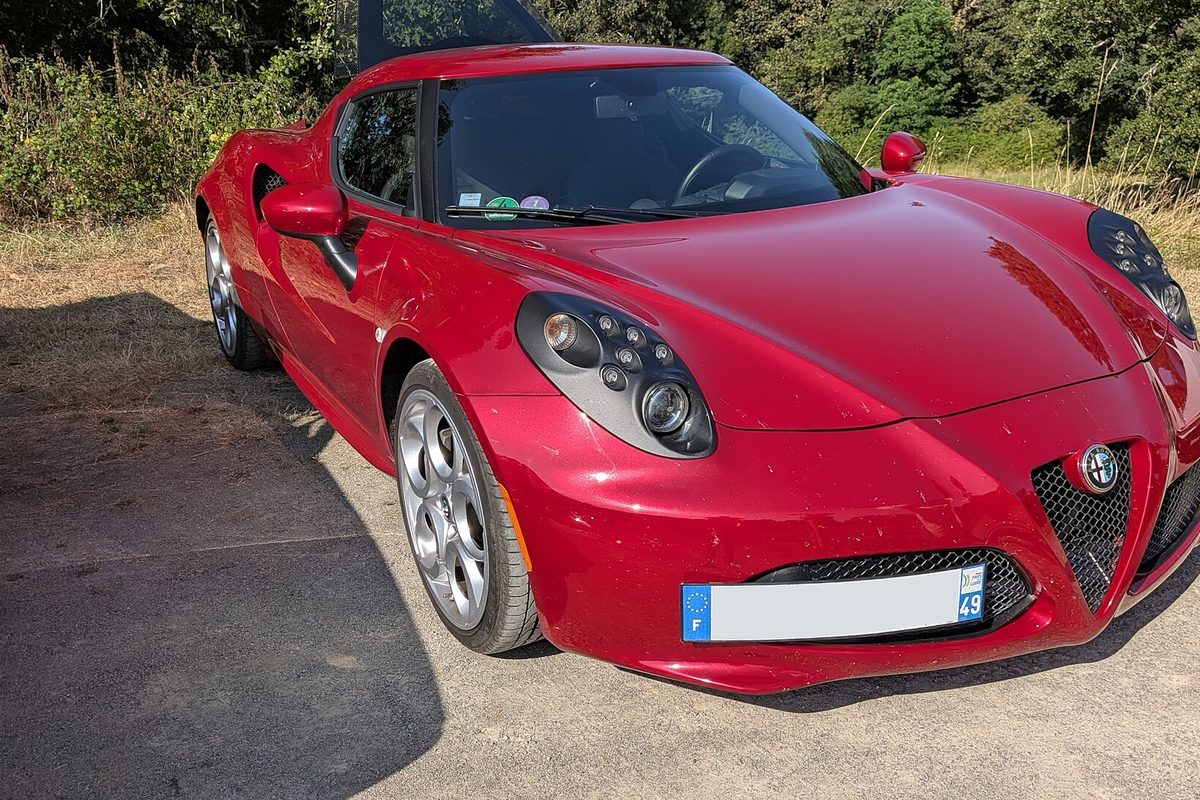 A red Alfa Romeo 4C coupe seen from the front corner, parked on gravel beside dry brush