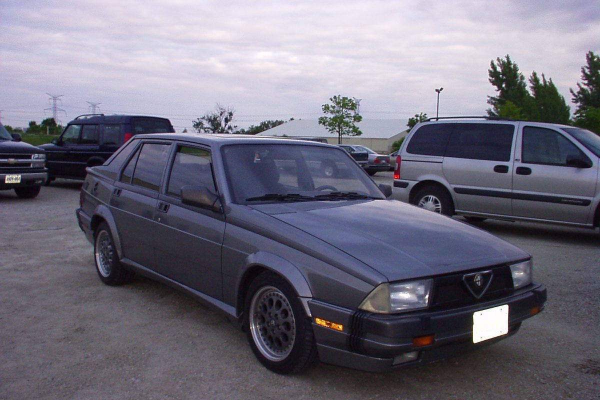 A grey Alfa Romeo Milano four-door sedan with alloy wheels, parked on a gravel lot beside a minivan
