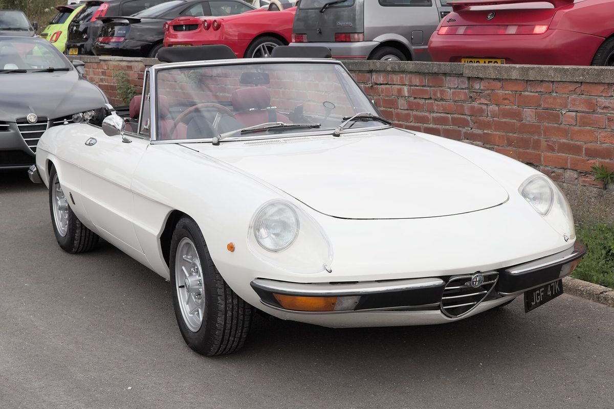 A white Alfa Romeo Spider convertible with its top down, chrome bumpers and round headlights, parked by a low brick wall