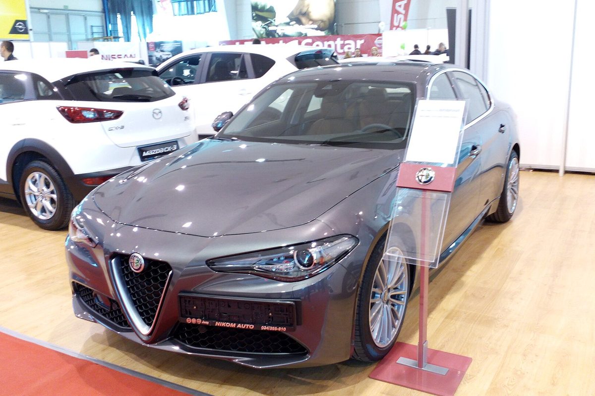 A gray Alfa Romeo Giulia sedan of the current generation parked on the floor of an indoor car show, with other cars on display behind it
