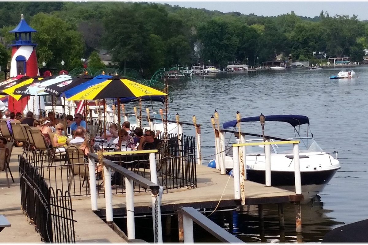 Diners under striped umbrellas on the waterfront patio at Port Edwards in Algonquin, with a small red and white lighthouse and a boat tied up at the dock