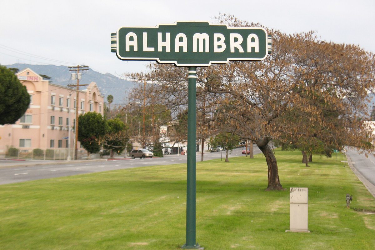 A green and white Alhambra welcome sign on a grassy median, with a leafless tree, a pink building and the San Gabriel Mountains behind