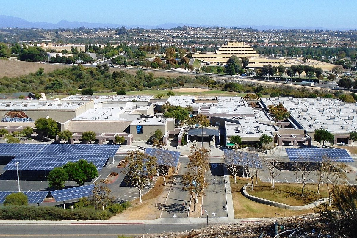 Aliso Niguel High School's flat roofs and solar-panel parking canopies seen from Hillview Park, with the terraced Chet Holifield Federal Building and hills behind