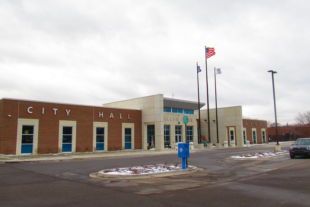 Allen Park City Hall, a brick and pale stone building with CITY HALL in large letters, flags out front and a dusting of snow on the curbs