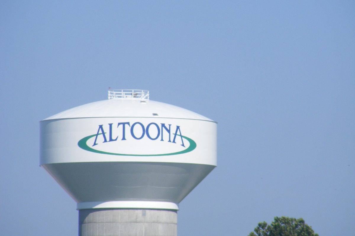 The top of a white water tower with ALTOONA in blue letters inside a green oval, against a clear blue sky