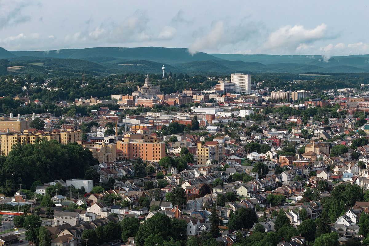 Aerial view over Altoona's rows of houses and brick buildings toward a domed church and a white high-rise, with long green ridges and clouds behind