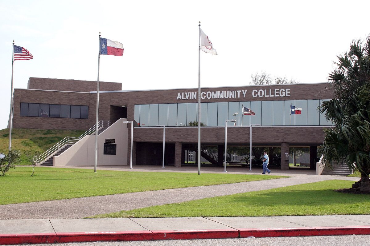 The brick main building of Alvin Community College with U.S., Texas and college flags on tall poles over a green lawn and a palm tree at the edge