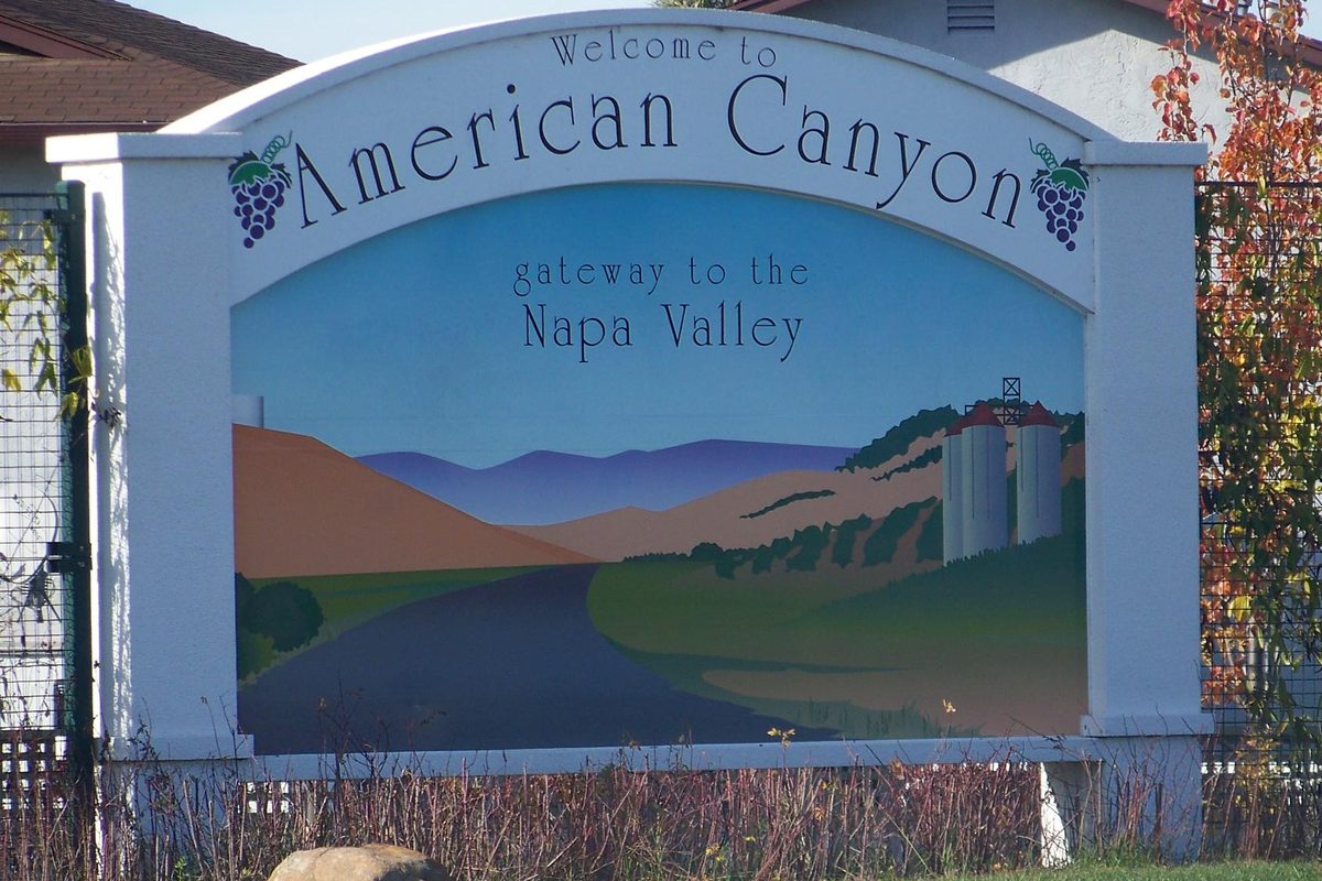 The Welcome to American Canyon sign reading Gateway to the Napa Valley, painted with hills, a road and grain silos, beside autumn shrubs