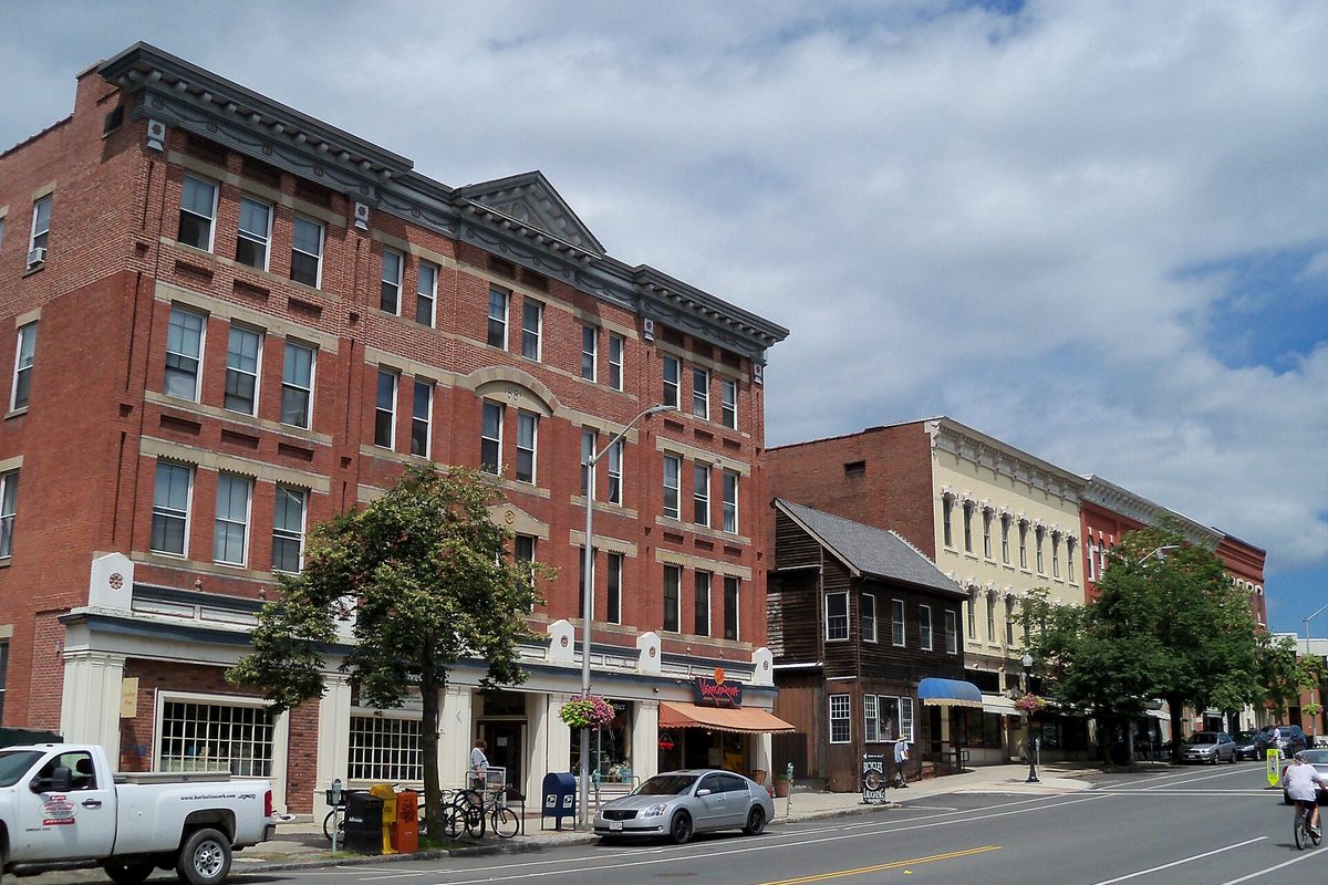 A four-story red brick building with shops at street level in downtown Amherst, a white pickup and a silver sedan parked at the curb and a cyclist riding past