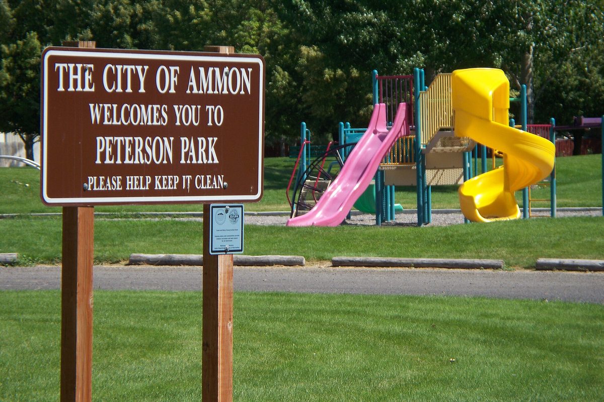A brown wooden sign reading The City of Ammon Welcomes You to Peterson Park, in front of a playground with pink and yellow slides