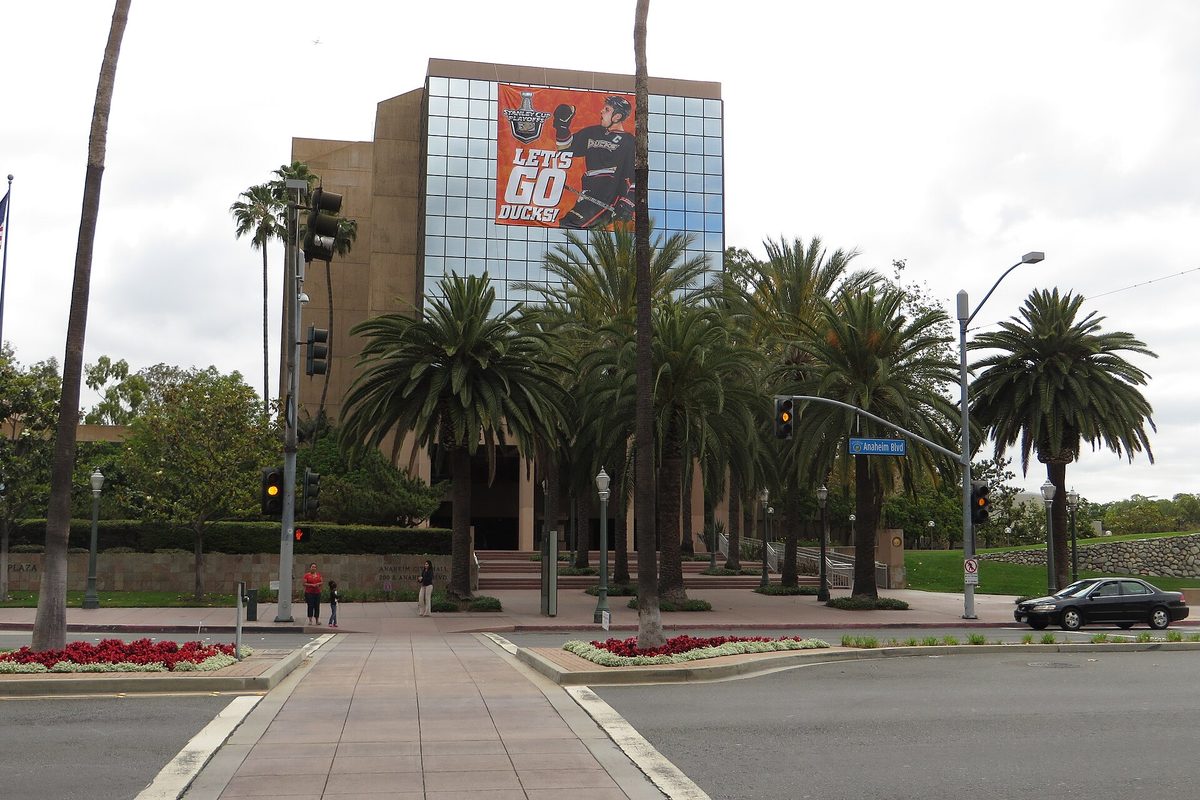 Anaheim City Hall, a glass and stone building with a Let's Go Ducks banner, behind tall palm trees, a red flower bed and a crosswalk