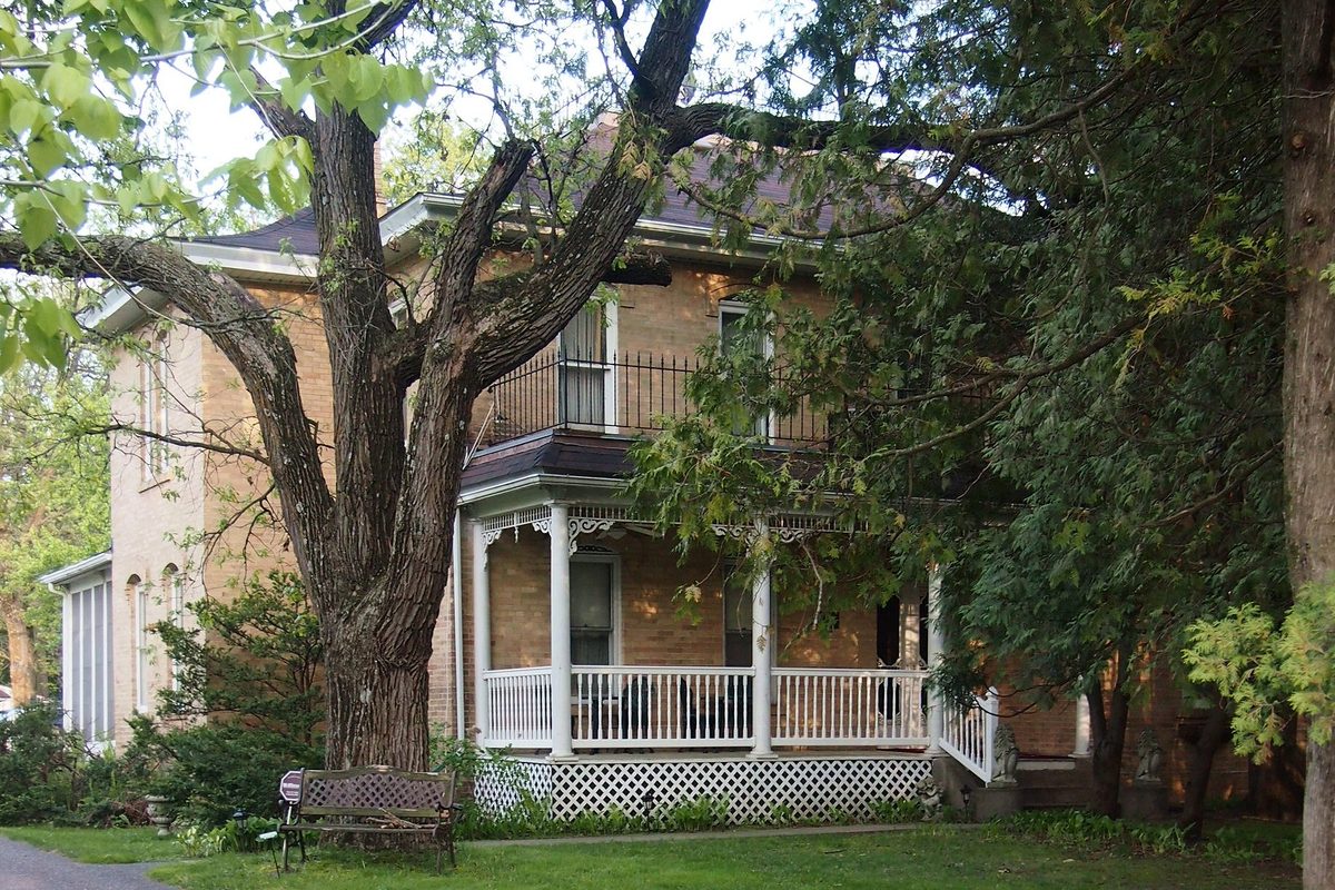 The brick Porter Kelsey House in Andover with a white wraparound porch and iron balcony, partly hidden behind a large old tree and green shrubs