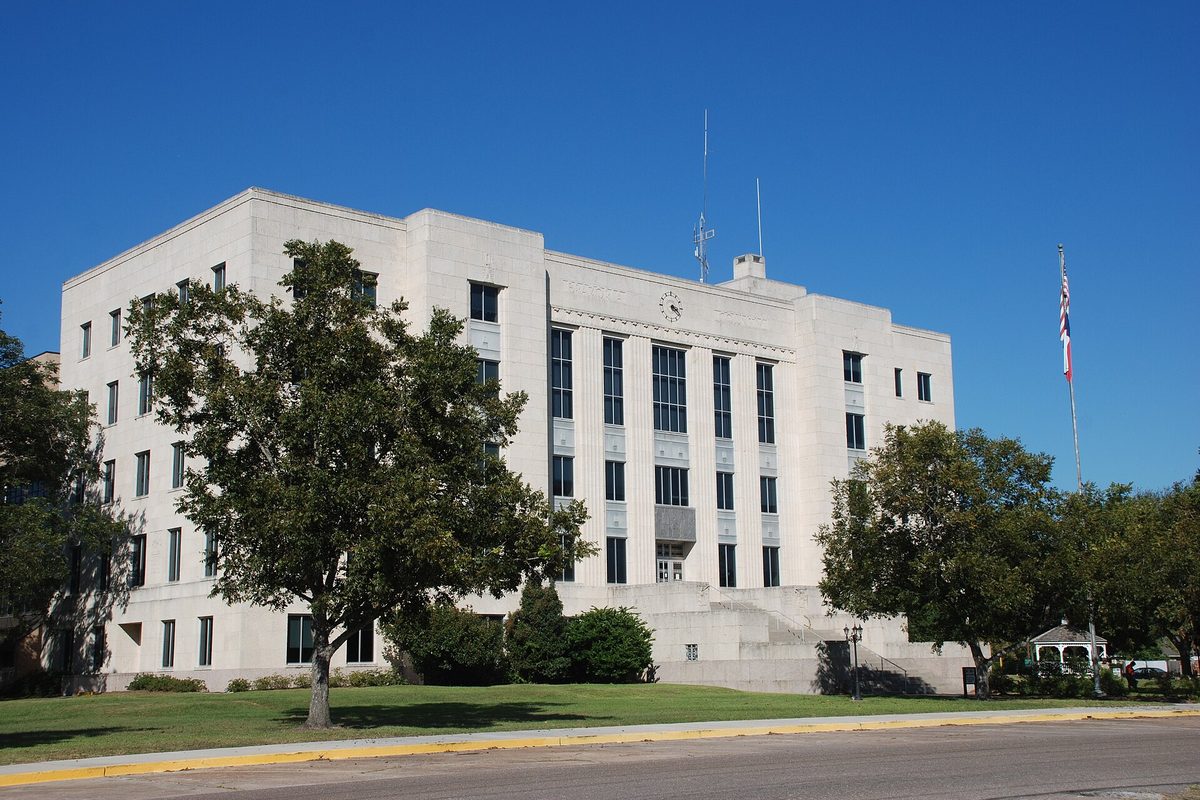 The white multistory Brazoria County Courthouse in Angleton, with tall vertical windows, a clock, a flagpole and large shade trees on the lawn