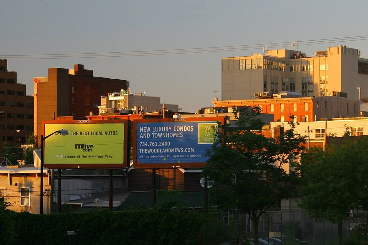 Two billboards in front of brick and pale concrete buildings in downtown Ann Arbor, lit orange by low evening sun