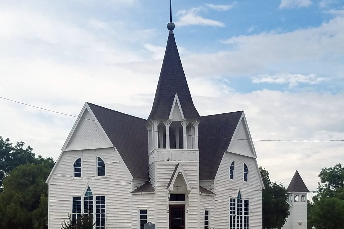 A white wooden church in Anna with a tall dark pointed steeple, arched windows and a smaller white bell tower behind it, under a partly cloudy sky