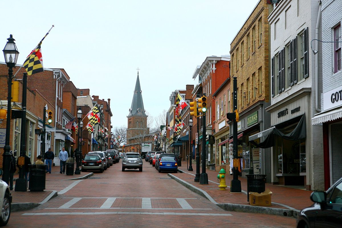 Brick-paved Main Street in downtown Annapolis lined with shops, Maryland flags on the lampposts and a church steeple at the far end on an overcast day