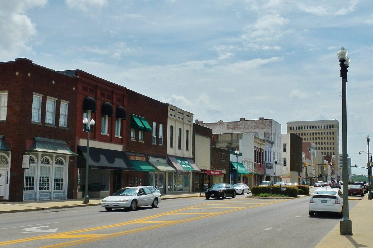 Downtown Anniston's main street lined with brick storefronts, green awnings and parked cars, with a tall office building at the far end