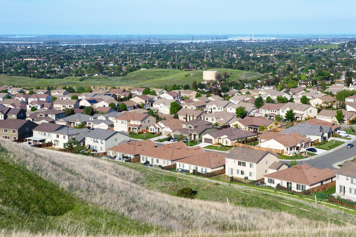Rows of tile-roofed Antioch homes seen from a dry grassy hillside, with the river and its bridge faint on the horizon