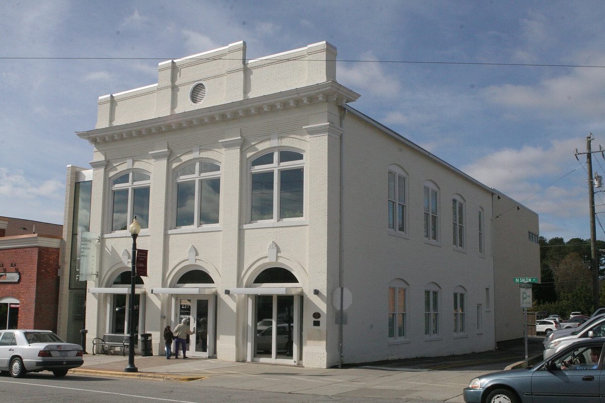 The white two-story historic Apex Town Hall with arched windows and a parapet, on a downtown street corner with parked cars