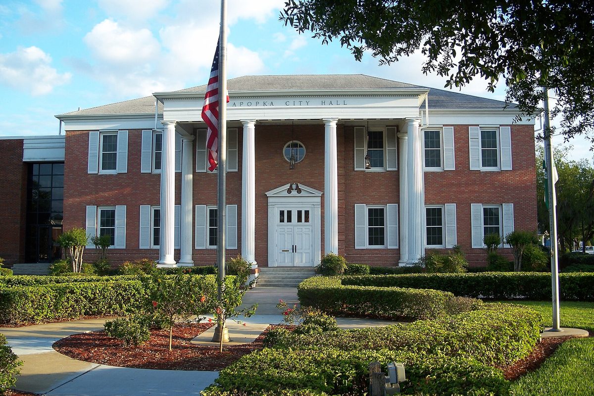 Apopka City Hall, a red brick building with a white-columned portico and an American flag, behind clipped hedges and a curved walk