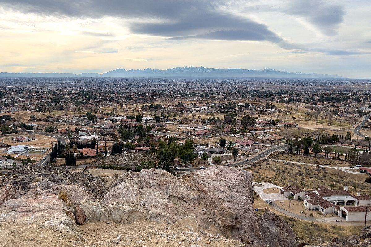 View southwest from the rocky top of Bass Hill over Apple Valley's spread-out homes and desert lots, with mountains on the horizon under a cloudy sky