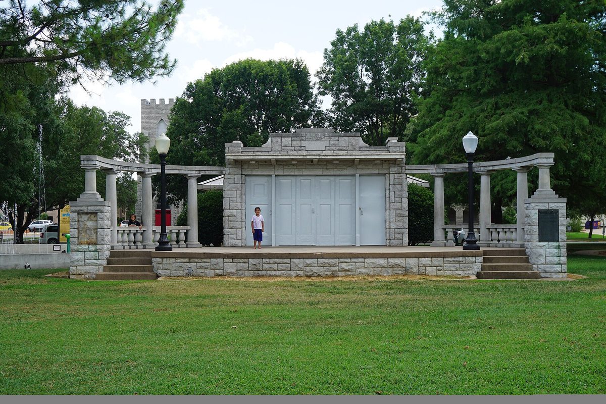 The stone bandstand in Ardmore's Central Park, with columned wings on each side, a child standing on the stage and a castle-style tower behind the trees