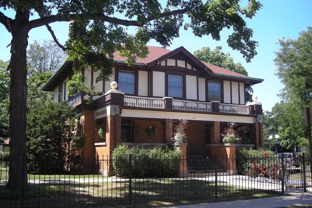 The Nathaniel Moore Banta House in Arlington Heights, a brick and stucco home with a wide front porch and an upper balcony, behind an iron fence and a big shade tree