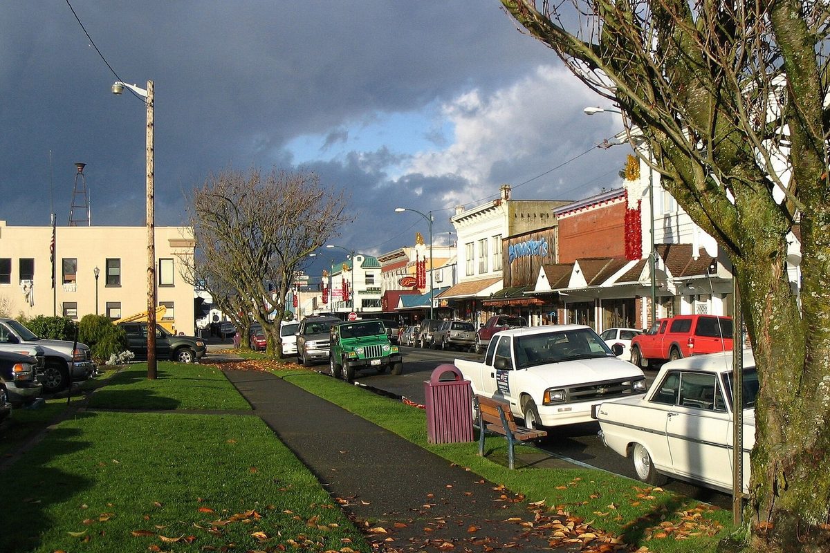 Downtown Arlington, Washington, with brick and wood storefronts, pickup trucks and a green Jeep parked along the curb, and autumn leaves on the grass under dark clouds